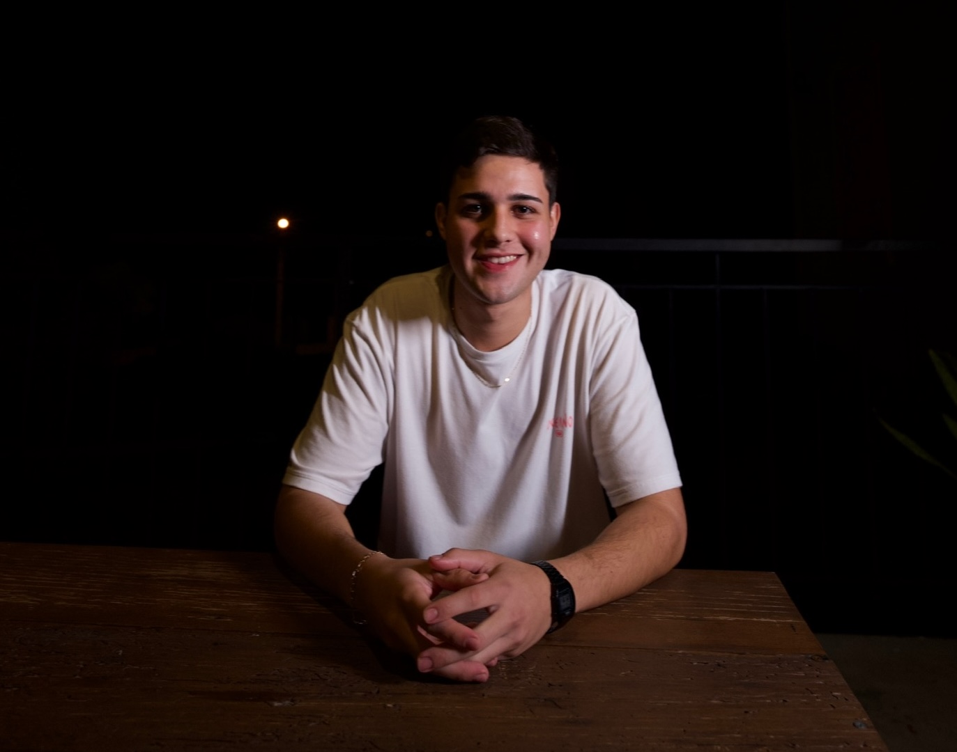 Foto de Daniel Nascimento, sorrindo, sentado em ambiente externo, fundo escuro, usando camiseta branca e relogio.